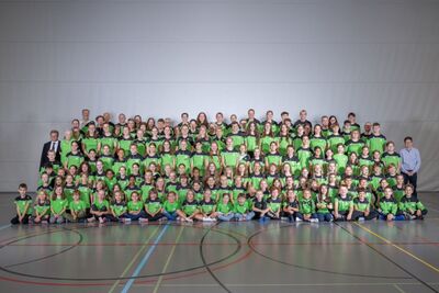 In einer grossen Gruppe posieren Mitglieder des Turnvereins aus Ingenbohl (6440, Kanton Schwyz) in grünen Vereinsshirts für ein gemeinsames Foto in der Sporthalle.