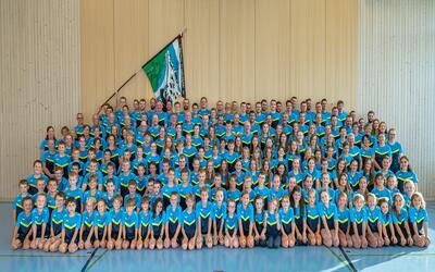 In einer Turnhalle in 9103 Schwellbrunn im Kanton Appenzell Ausserrhoden steht eine grosse Gruppe in einheitlichen blauen Sportshirts für ein Gruppenfoto zusammen.