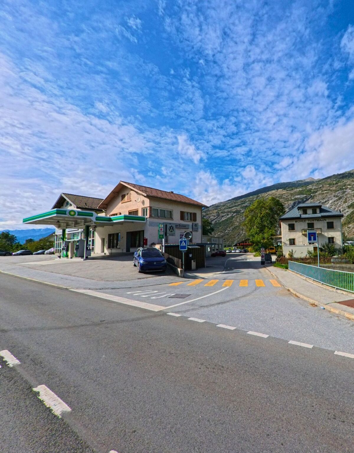 An einer Strasse in Turtmann-Unterems (3946, Kanton Wallis) steht eine Tankstelle mit Wohnhaus, daneben sind Fussgaengerstreifen und weitere Gebaeude sichtbar.