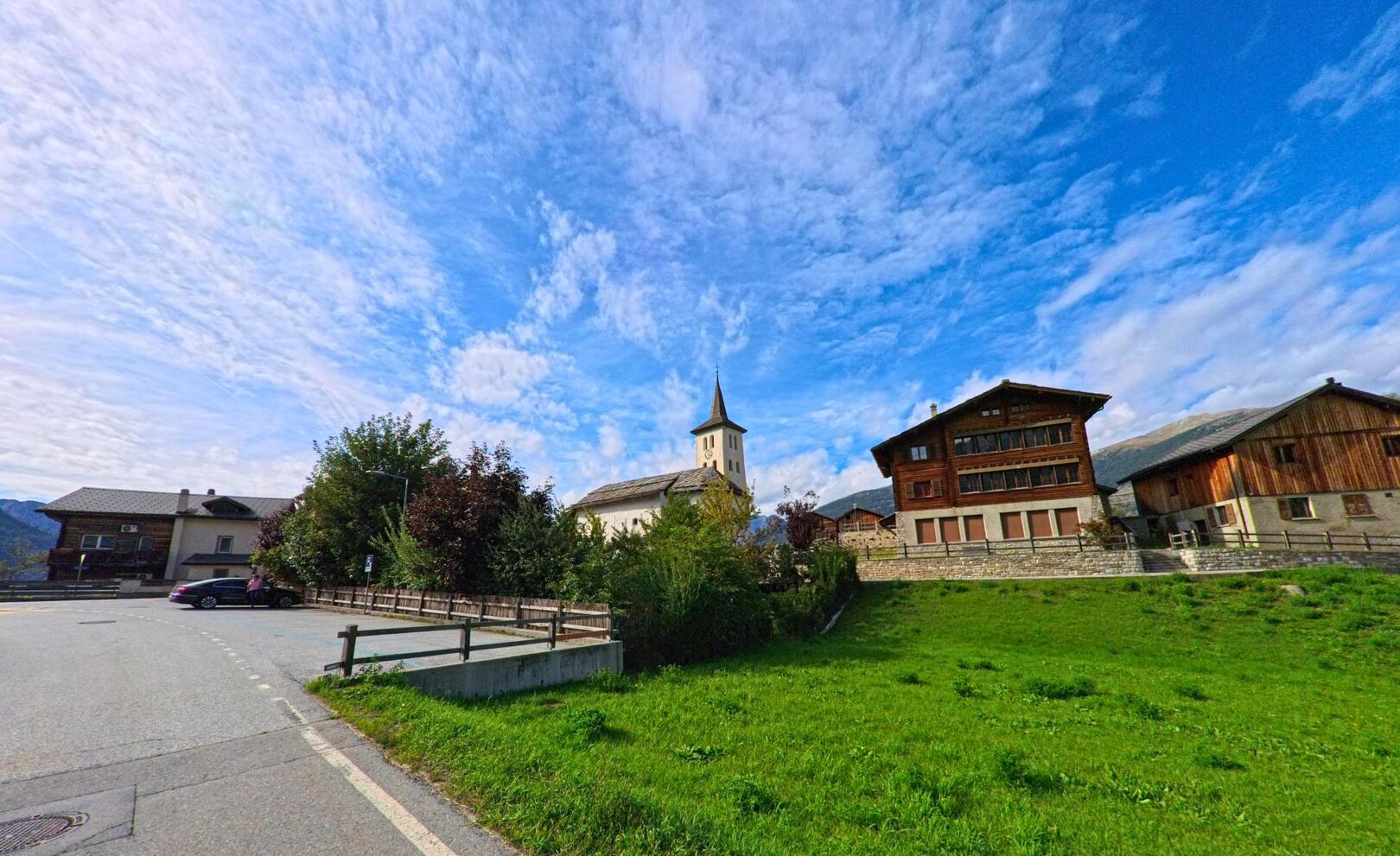 Die Kirche mit markantem Turm steht in Turtmann-Unterems (3948, Kanton Wallis) zwischen traditionellen Wohnhaeusern und gruener Wiese.