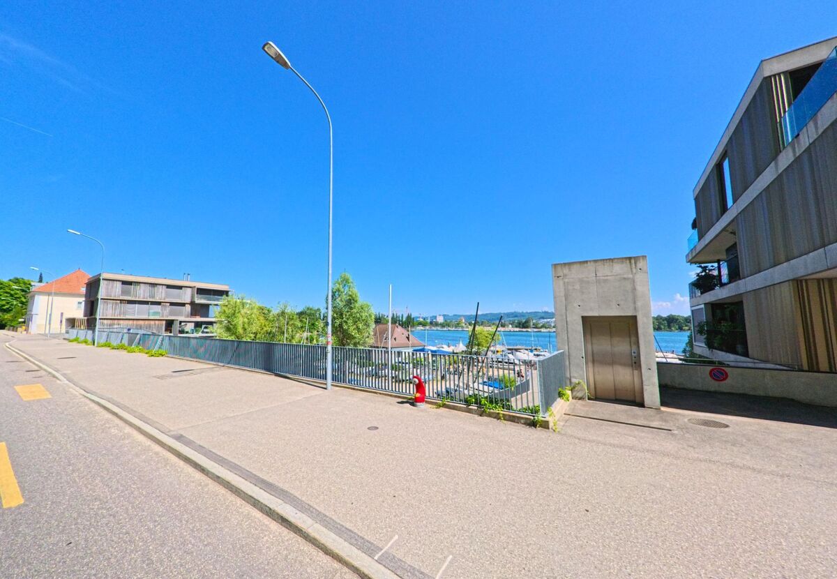 In Twann (2505, Kanton Bern) führt die Straße entlang moderner Wohngebäude mit Blick auf den See und die Yachten, umrahmt von klarem blauen Himmel.