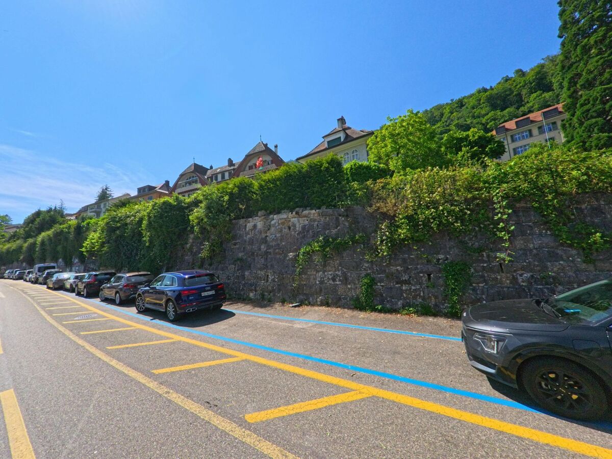 In Twann (2505, Kanton Bern) verläuft die Straße entlang einer Parkzone, die von üppigem Grün und historischen Gebäuden überragt wird, mit einem klaren blauen Himmel im Hintergrund.