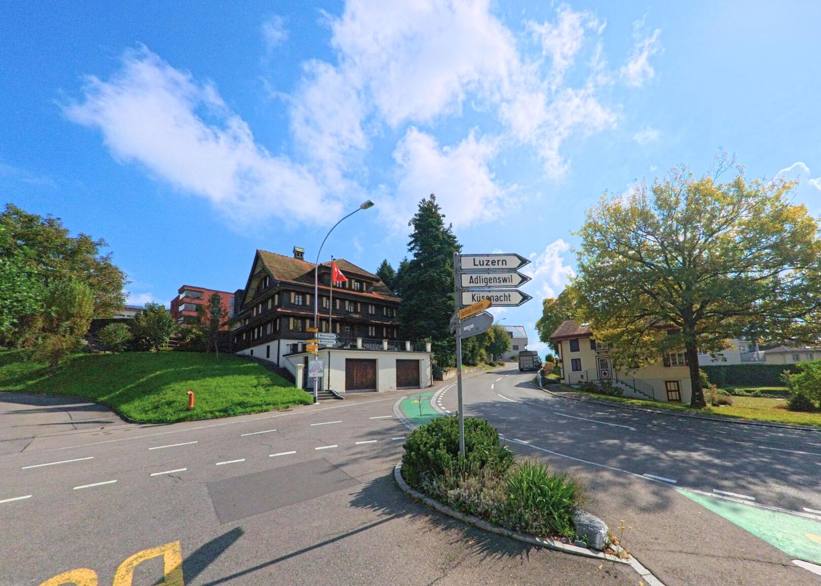 In Udligenswil (6044, Kanton Luzern) treffen mehrere Strassen an einer Kreuzung zusammen, umgeben von einem traditionellen Holzhaus am Hang und Wegweisern vor einem klaren Himmel.