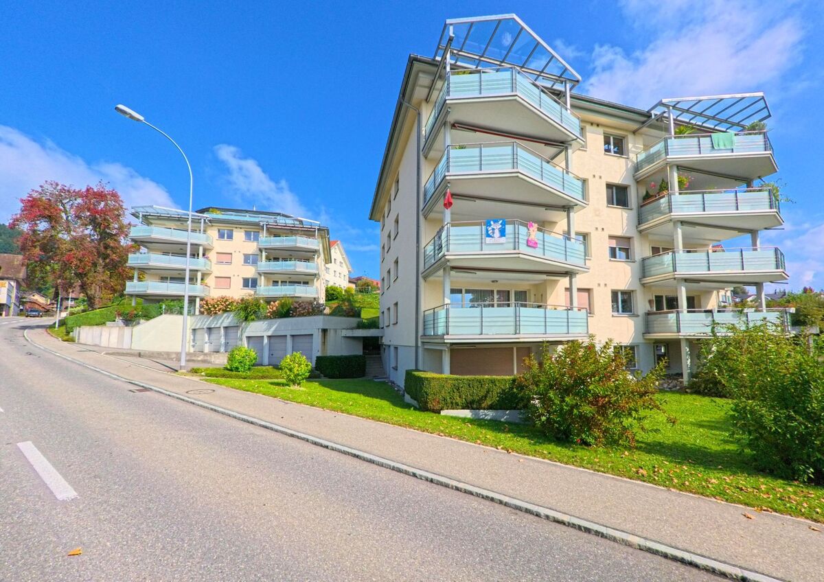 In Udligenswil (6044, Kanton Luzern) stehen moderne Mehrfamilienhaeuser mit grossen Balkonen an einer leicht ansteigenden Strasse, eingerahmt von Gruenflaechen und einem klaren Himmel.