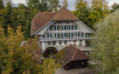 Eine historische Muehle mit Wasserrad liegt umgeben von Baeumen in Luetzelflueh (3432, Kanton Bern) und zeigt ein idyllisches Gebaeude.