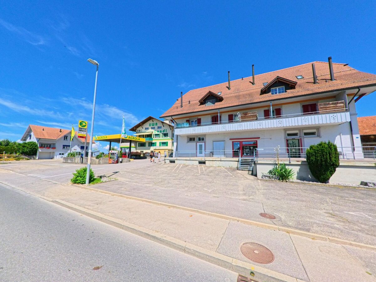  In Ueberstorf (3182, Kanton Freiburg) liegt gegenüber einer Bushaltestelle ein grosses Gebäude mit Ladenlokalen neben einer Agrola-Tankstelle.