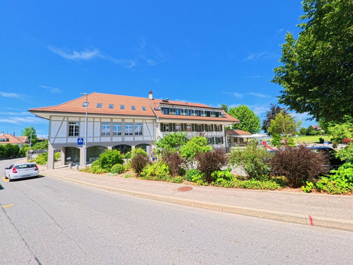 In Ueberstorf (3182, Kanton Freiburg) steht ein grosses Gebäude mit rotem Ziegeldach und begrüntem Vorplatz an einer ruhigen Strasse.