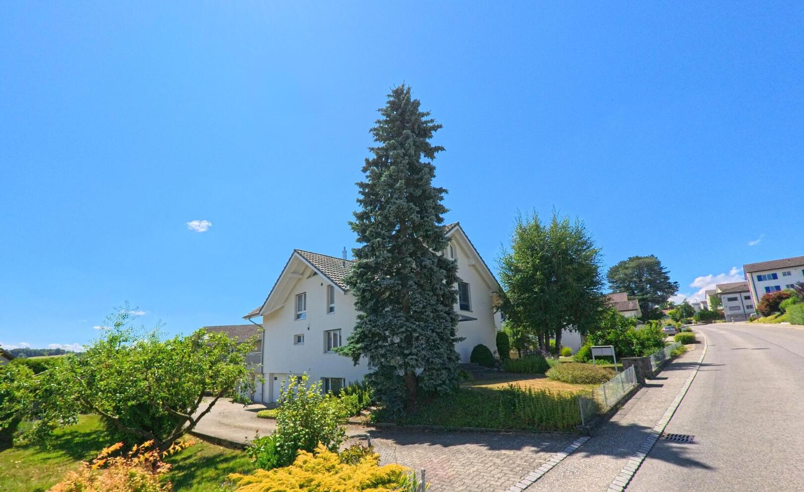 An einer leicht ansteigenden Strasse in Ueberstorf (3182, Kanton Freiburg) steht ein weiss gestrichenes Wohnhaus mit grossem Vorgarten und hohen Bäumen.