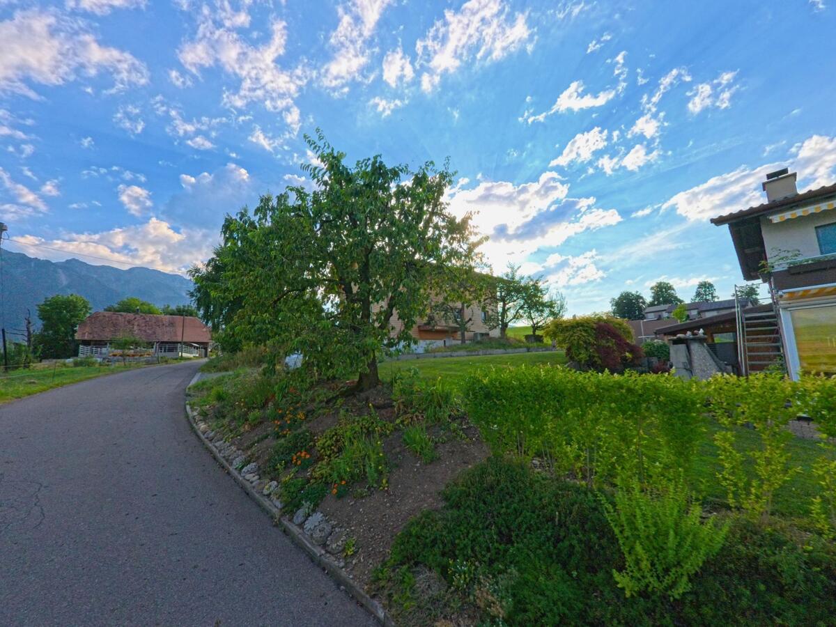 In Uebeschi (3635, Kanton Bern) verläuft eine schmale Strasse an einem blühenden Vorgarten mit Baum vorbei, flankiert von Bauernhäusern und modernen Wohnbauten.