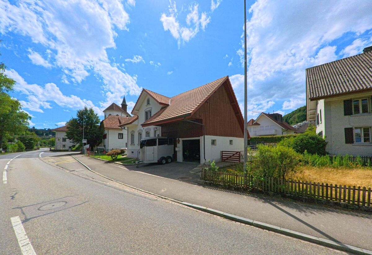 In Uerkheim (4813, Kanton Aargau) verläuft eine leicht kurvige Strasse an einem Bauernhaus mit angebautem Unterstand vorbei, im Hintergrund ist der Kirchturm des Orts sichtbar.