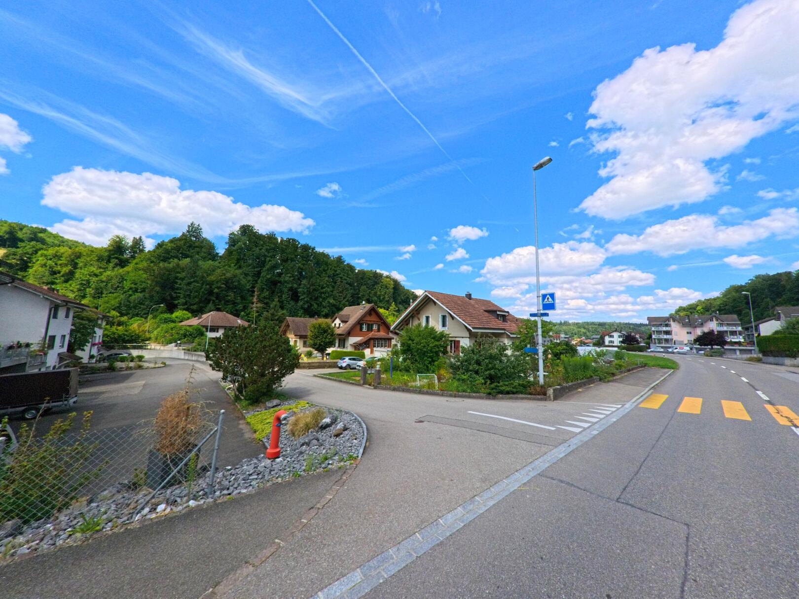 In Uerkheim (4813, Kanton Aargau) zweigt eine Nebenstrasse mit gepflegten Einfamilienhäusern von der Hauptstrasse ab, während ein Zebrastreifen und ein Fussgängerschild den Übergang markieren.