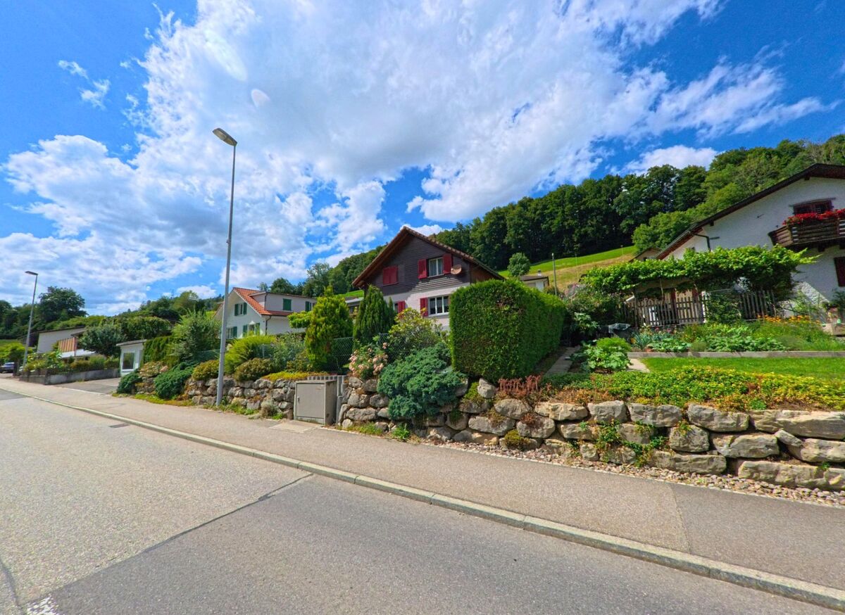 In Uerkheim (4813, Kanton Aargau) stehen entlang einer Wohnstrasse gepflegte Einfamilienhäuser mit farbenfrohen Gärten und Trockensteinmauern vor einem bewaldeten Hang.