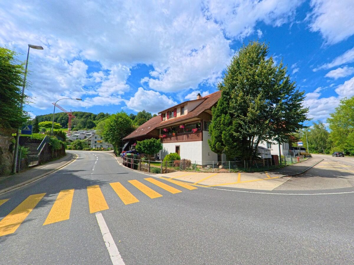 In Uerkheim (4813, Kanton Aargau) treffen sich zwei Strassen an einer Kreuzung mit Fussgängerstreifen, flankiert von einem traditionellen Haus mit Blumenbalkon und einem Treppenaufgang.