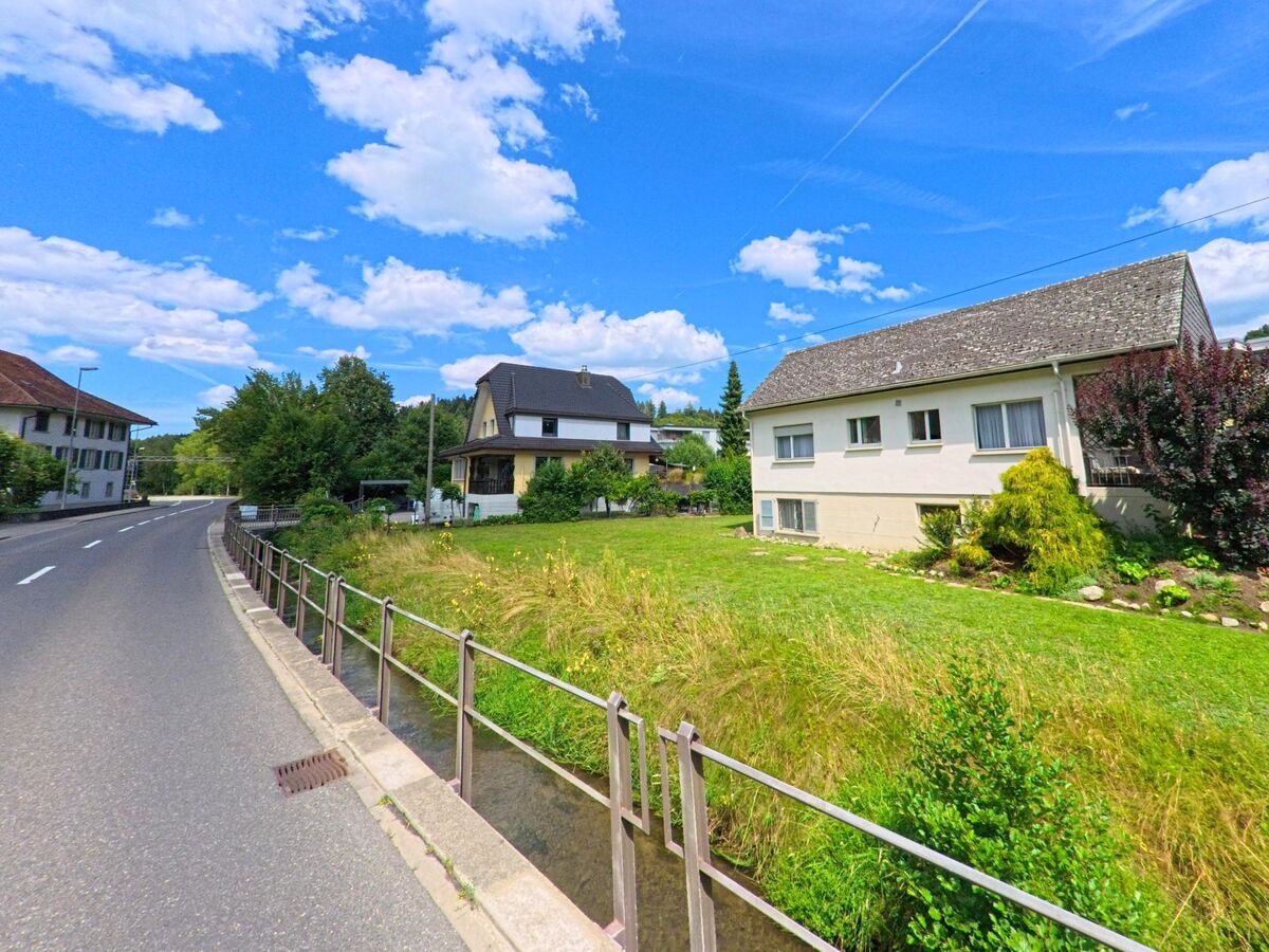 In Uerkheim (4813, Kanton Aargau) verläuft eine schmale Strasse mit Geländer entlang eines Bachs, flankiert von Einfamilienhäusern mit gepflegten Gärten und Wiese.