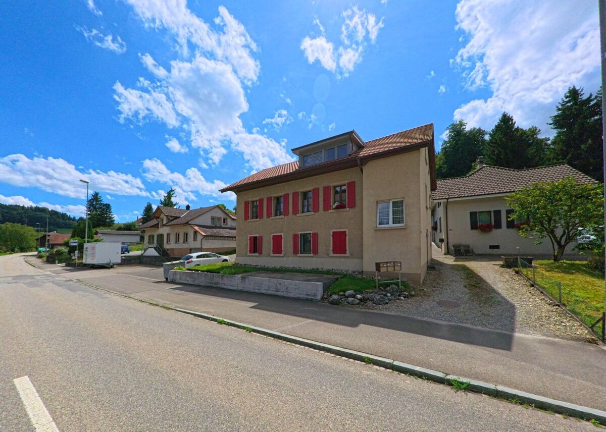 In Uerkheim (4813, Kanton Aargau) säumen Wohnhäuser mit roten Fensterläden eine leicht ansteigende Hauptstrasse, im Hintergrund führen Zufahrten zu weiteren Liegenschaften.