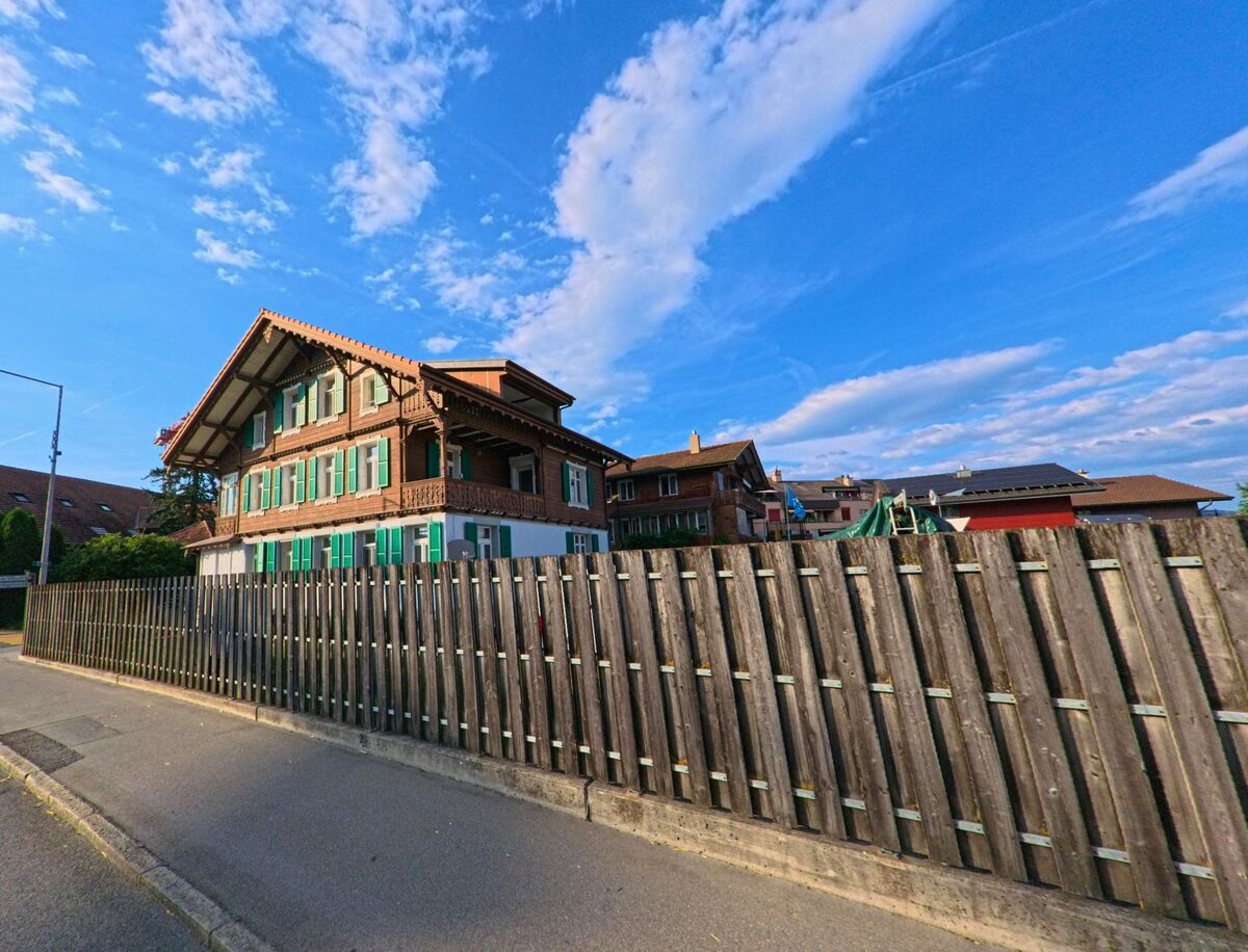 Ein traditionelles Berner Wohnhaus mit grünen Fensterläden und holzverzierter Fassade steht in Uetendorf (3661, Kanton Bern) hinter einem hohen Holzzaun.