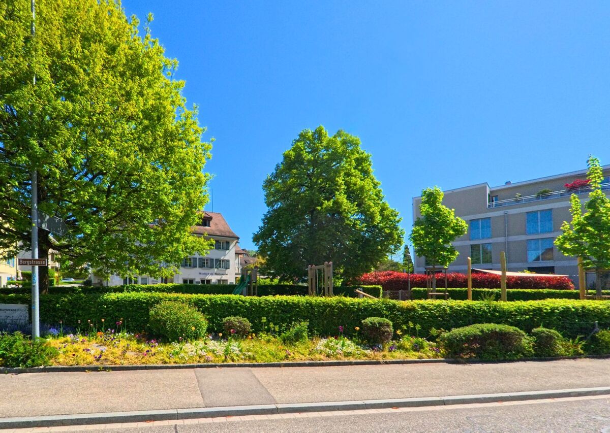 In Uetikon am See (8707, Kanton Zürich) säumen bunte Blumenbeete und dichte Hecken einen Bereich mit Bäumen und einem Spielplatz nahe der Bergstrasse.