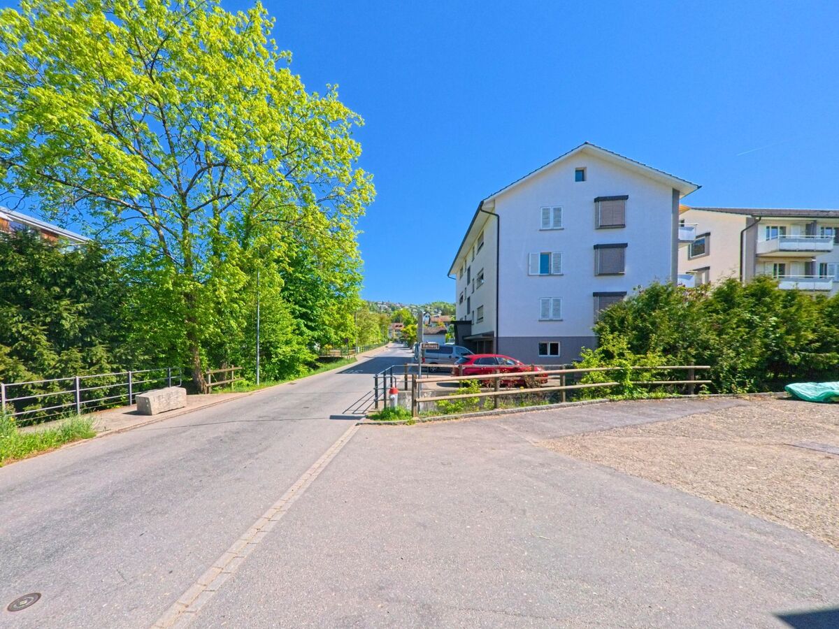 In Uetikon am See (8707, Kanton Zürich) führt eine schmale Strasse mit Gehwegbrücke an Bäumen und Wohnhäusern vorbei in ein ruhiges Quartier.