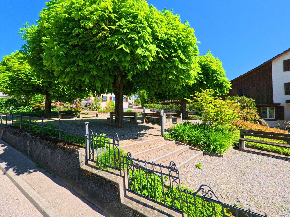 Ein kleiner Platz mit Sitzbänken und schattenspendenden Kastanienbäumen liegt ruhig zwischen Wohnhäusern in Uetikon am See (8707, Kanton Zürich).