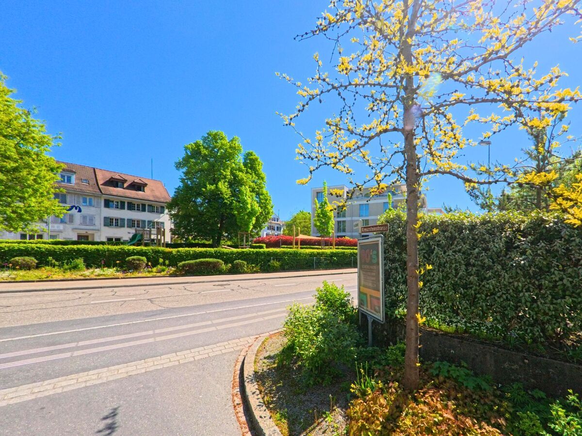 In Uetikon am See (8707, Kanton Zürich) verläuft eine Strasse mit Gleisen entlang gepflegter Hecken und blühender Bäume, gegenüber liegt ein Wohnquartier mit Spielplatz.
