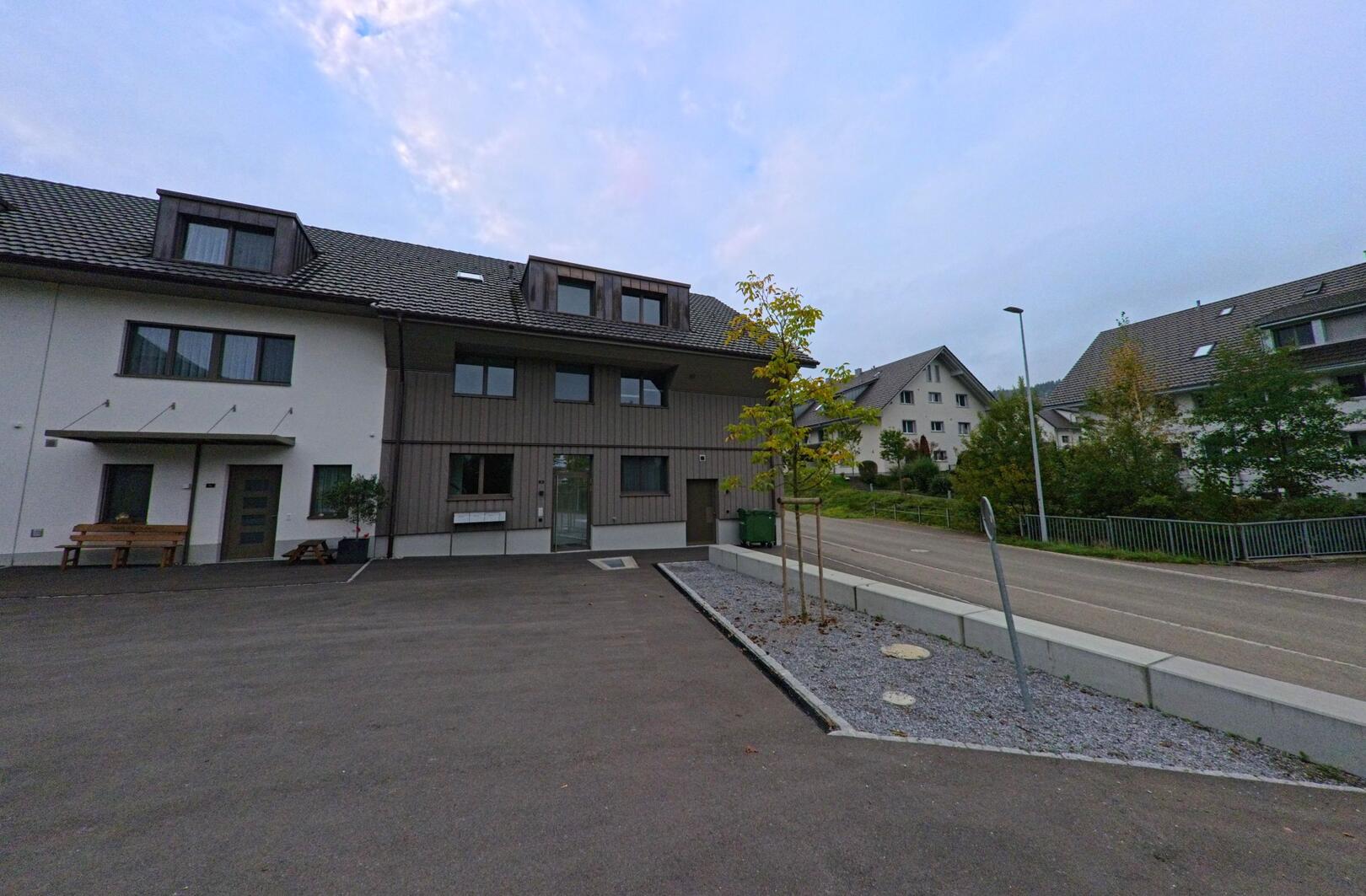In einer ruhigen Wohnstrasse von Uezwil (5619, Kanton Aargau) stehen moderne Mehrfamilienhaeuser mit frisch gepflanzten jungen Baeumen neben einer breiten Einfahrt.