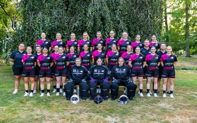 Die Mitglieder von UH Lejon Zäziwil aus Konolfingen (3510, Kanton Bern) posieren gemeinsam in schwarz-pinkfarbener Vereinskleidung mit drei Torhüterinnen in Schutzausrüstung für ein Gruppenfoto im Grünen.