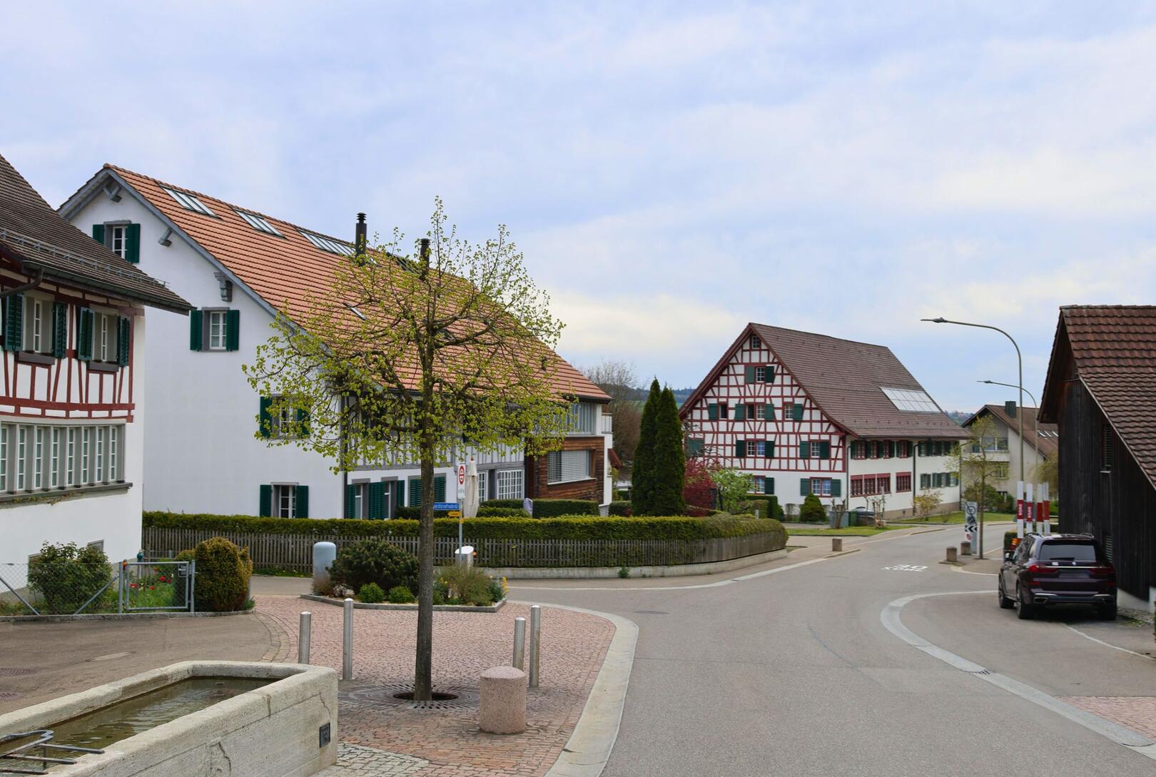 In der Dorfmitte von Uitikon (8142, Kanton Zürich) stehen mehrere gut erhaltene Fachwerkhäuser entlang einer ruhigen Strasse, im Vordergrund ein kleiner Brunnen mit Baum.