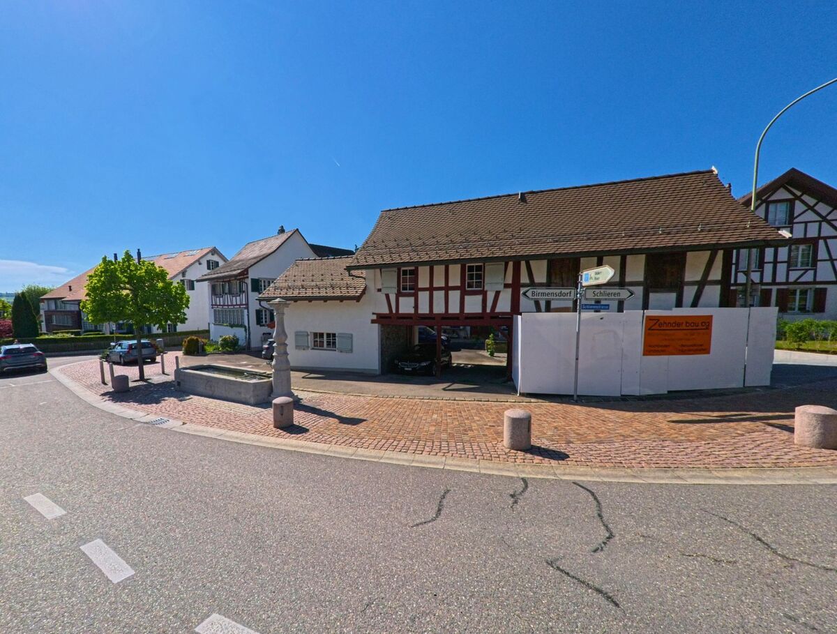 In Uitikon (8142, Kanton Zürich) steht an einer kleinen Platzanlage mit Brunnen ein traditionelles Fachwerkhaus, daneben zeigen Wegweiser die Richtungen nach Birmensdorf und Schlieren.