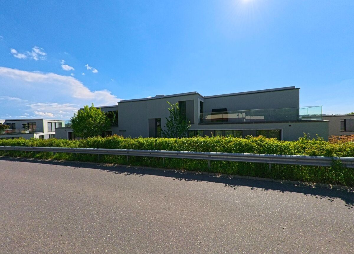 Hinter einer grünen Hecke stehen in Uitikon (8142, Kanton Zürich) moderne Flachdachbauten mit grossen Glasbalkonen an einer Strasse mit Leitplanke.