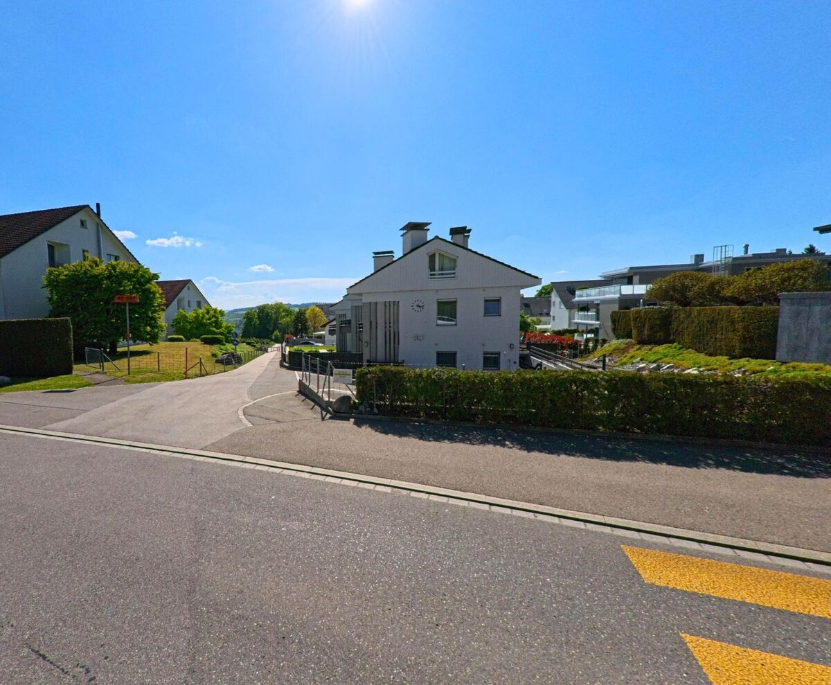 In Uitikon (8142, Kanton Zürich) führt eine Nebenstrasse an einem kleinen weissen Wohnhaus mit Uhr an der Fassade vorbei, umgeben von gepflegten Gärten.