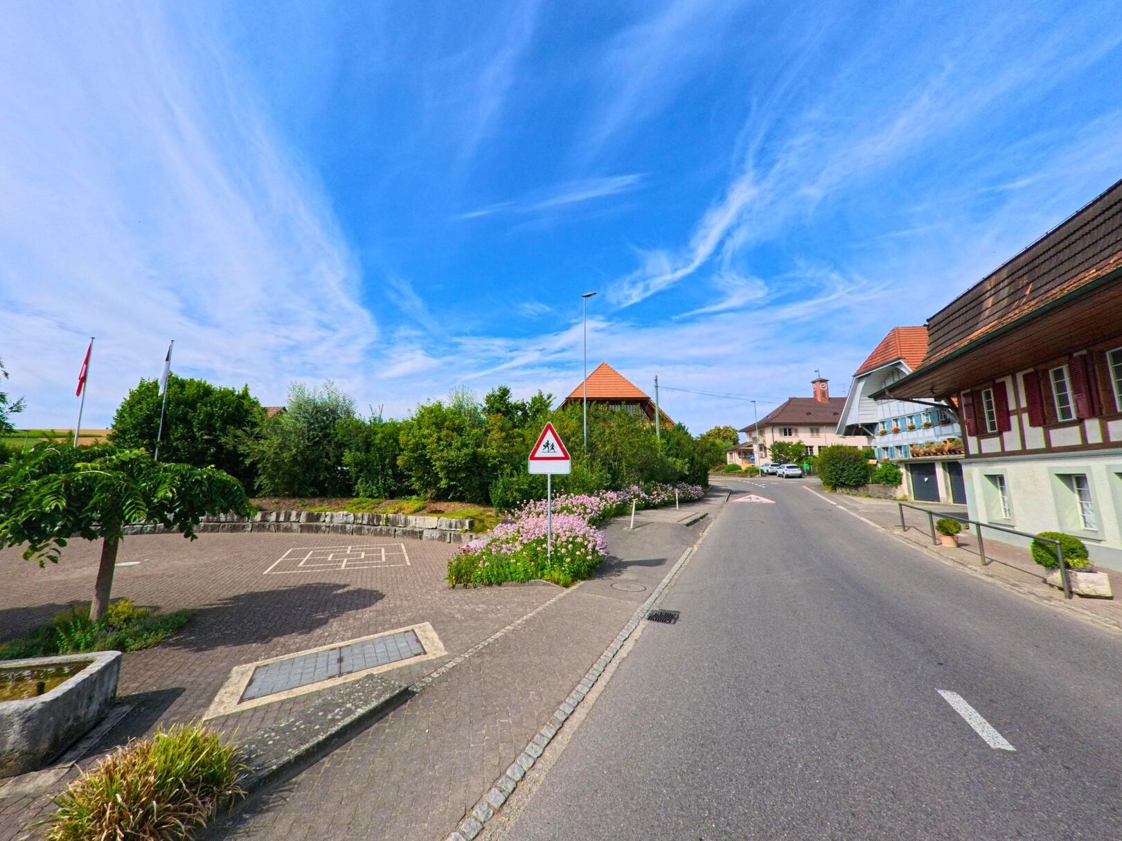 In Ulmiz (3214, Kanton Freiburg) verläuft eine ruhige Dorfstrasse mit gut erhaltenen Häusern und einem angrenzenden Schulareal unter klarem Himmel.