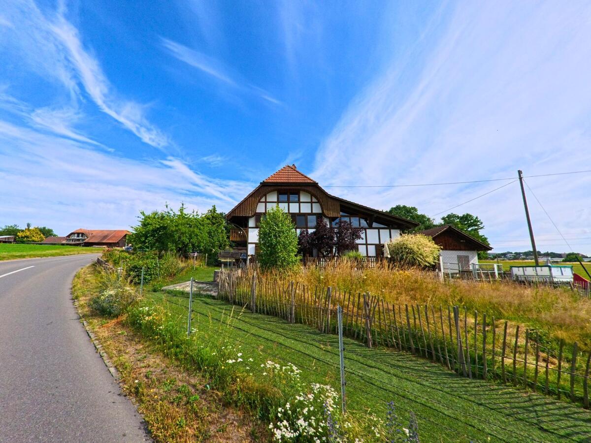 Ein typisches Bauernhaus mit Fachwerk steht in Ulmiz (3214, Kanton Freiburg) an einer leicht geschwungenen Landstrasse zwischen Feldern.