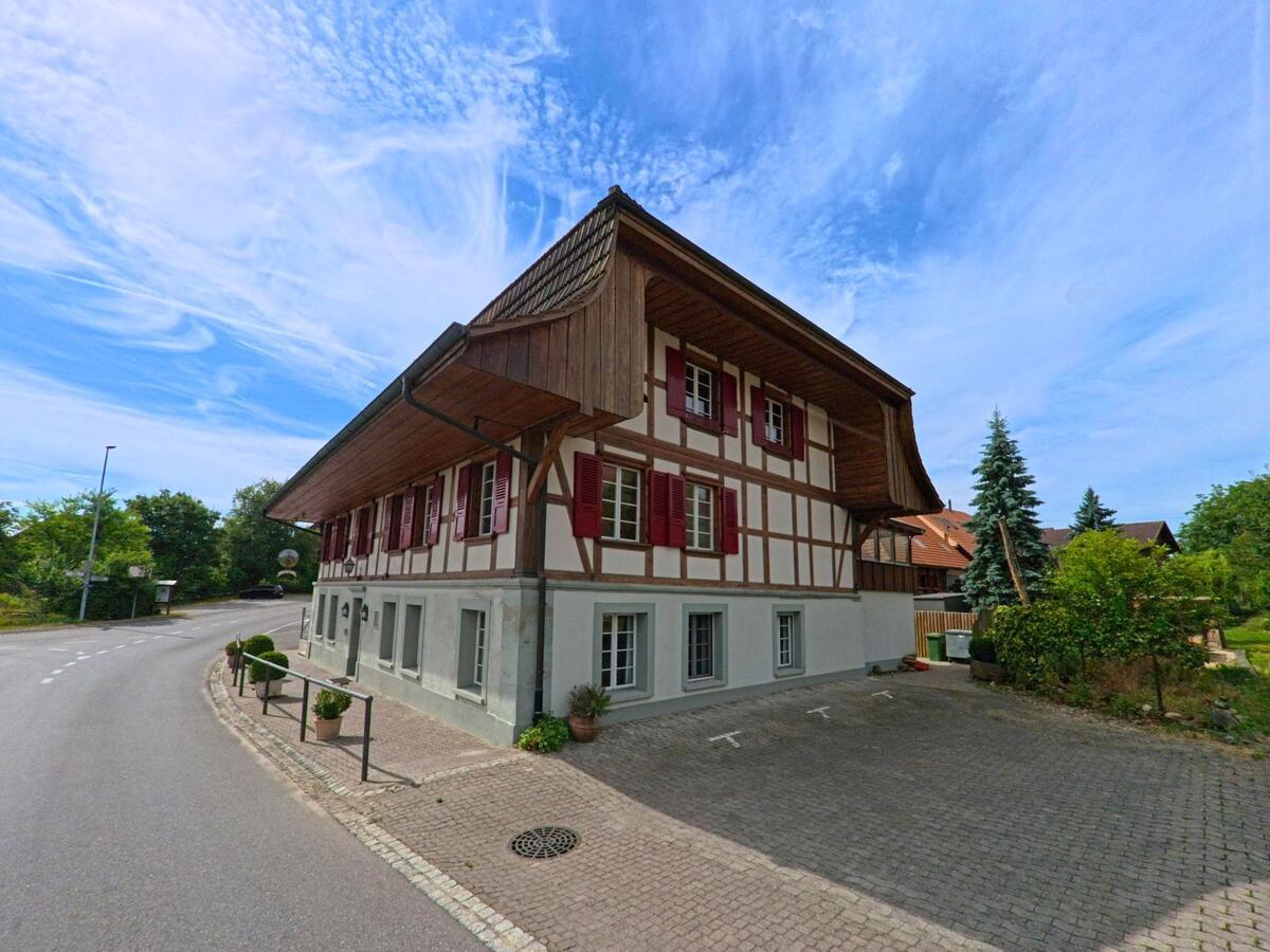 In Ulmiz (3214, Kanton Freiburg) steht ein gut erhaltenes Fachwerkhaus mit roten Fensterläden an einer Kurve der Hauptstrasse.