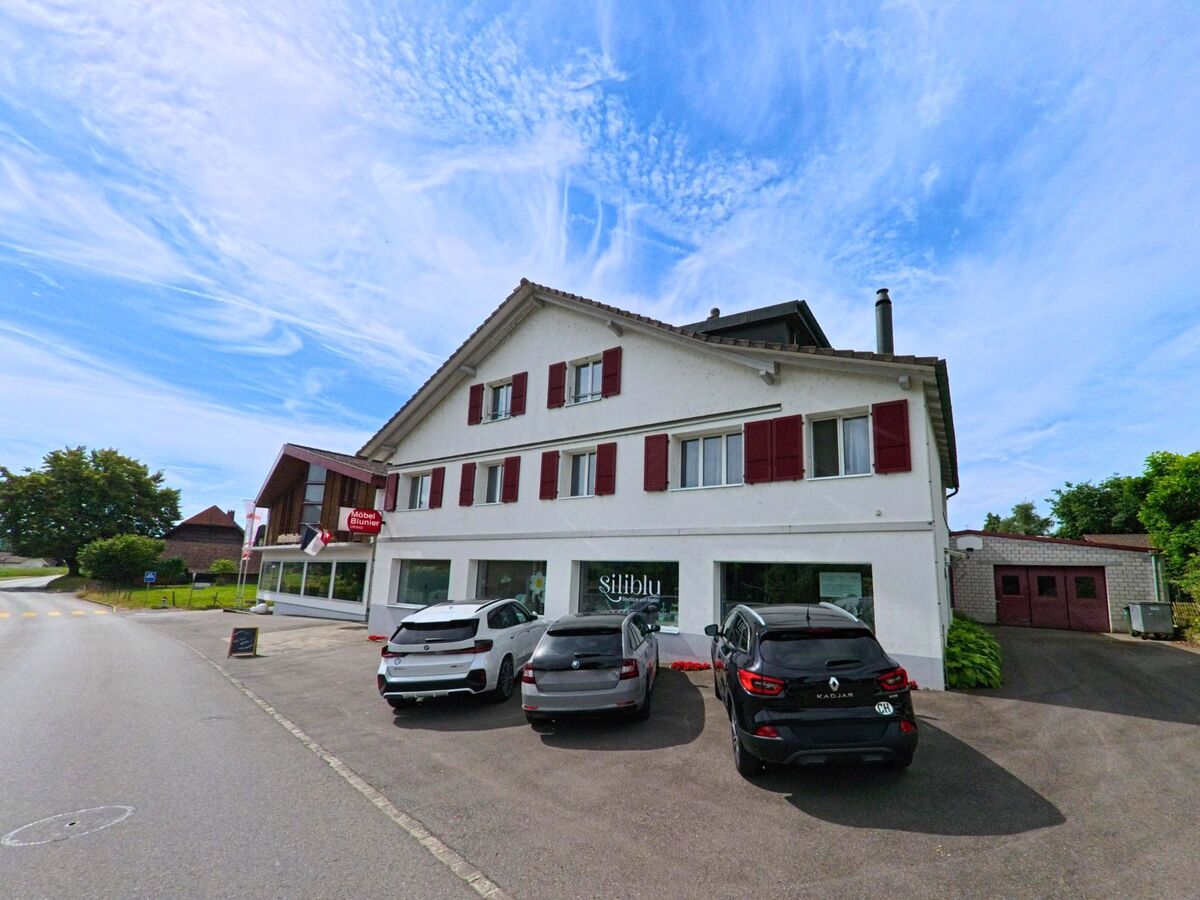 In Ulmiz (3214, Kanton Freiburg) befindet sich ein Geschäftshaus mit roten Fensterläden und einem Möbel- und Blumengeschäft direkt an der Hauptstrasse.