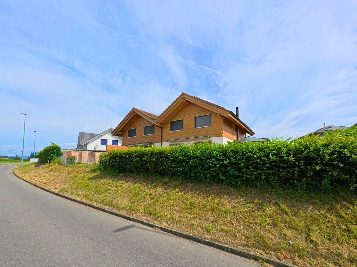 Am Ortsrand von Ulmiz (3214, Kanton Freiburg) steht ein modernes Einfamilienhaus mit Holzfassade an einer leicht geschwungenen Strasse.