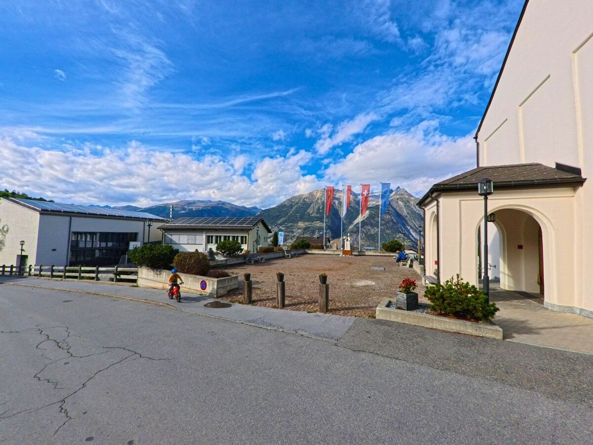 Vor der Kirche in Unterbaech (3944, Kanton Wallis) stehen mehrere Fahnen auf einem Platz mit Blick auf die umliegenden Berge.