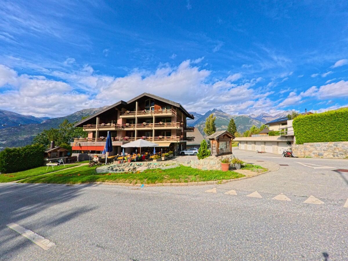 Ein mehrstoekiges Gebaeude mit Balkonen und Terrasse in Unterbaech (3944, Kanton Wallis) steht vor einer eindrucksvollen Bergkulisse, waehrend der Aussenbereich mit Sonnenschirmen gestaltet ist.