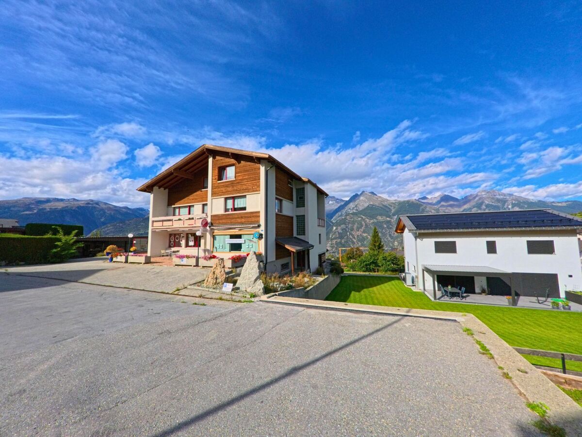 Ein modernes Wohnhaus mit Holzelementen und Balkonen steht in Unterbaech (3944, Kanton Wallis) neben einem Neubau mit Solardach, umgeben von gepflegten Gaerten und Bergkulisse.