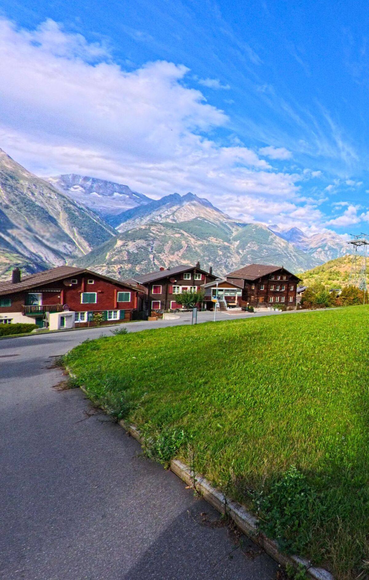 Vor der Kulisse hoher Berge im Kanton Wallis stehen in Unterbaech (3944) mehrere traditionelle Wohnhaeuser entlang einer Strasse neben einer gruene Wiese.