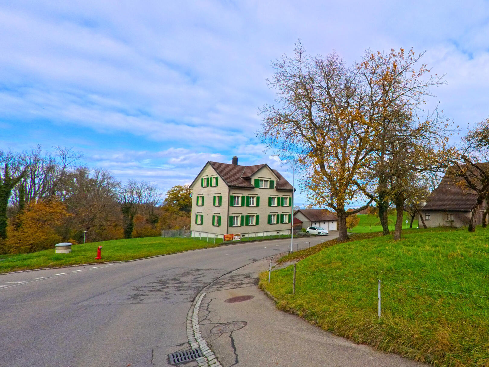 Ein Wohnhaus mit gruenen Fensterlaeden steht an einer leichten Strassenkurve in Untereggen (9303, Kanton St. Gallen) neben einer Wiese.