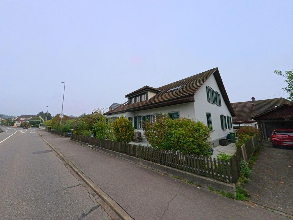 Ein Einfamilienhaus mit gruener Fensterlaeden steht in Unterentfelden (5035, Kanton Aargau) an einer leicht gekruemmten Strasse mit Gehweg und Vorgarten.