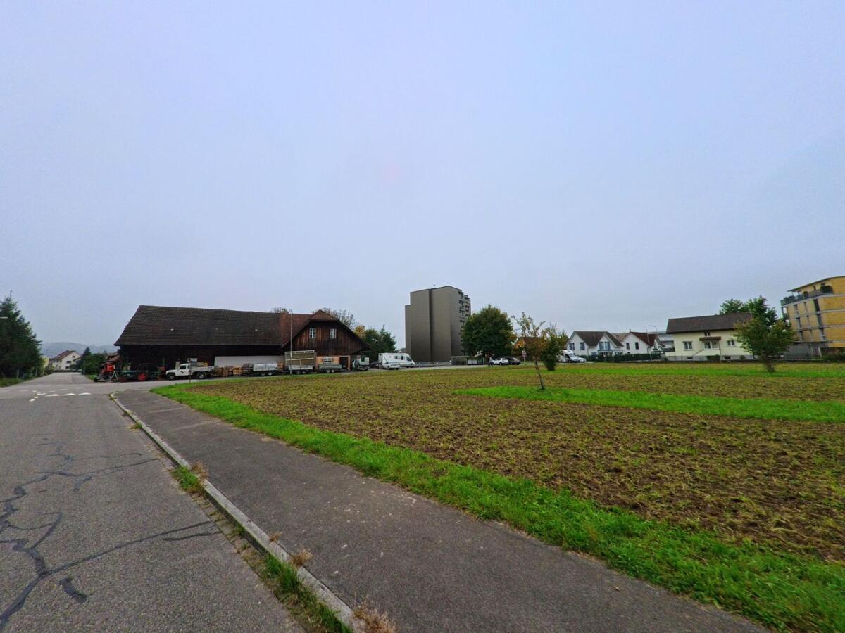 Am Rand von Unterentfelden (5035, Kanton Aargau) liegen ein landwirtschaftliches Gebaeude und offene Felder, dahinter stehen moderne und traditionelle Wohnhaeuser.