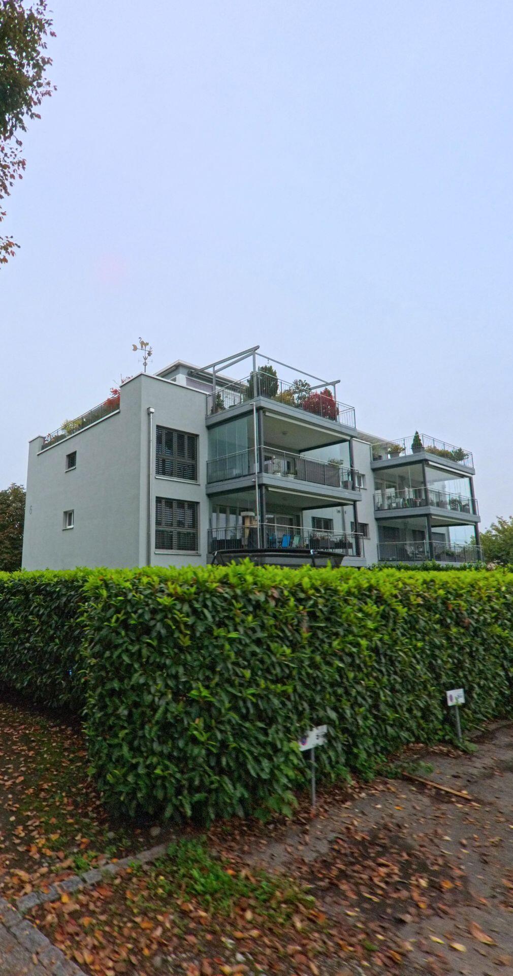 In Unterentfelden (5035, Kanton Aargau) steht ein modernes Mehrfamilienhaus mit grossen Balkonen hinter einer dichten gruene Hecke.