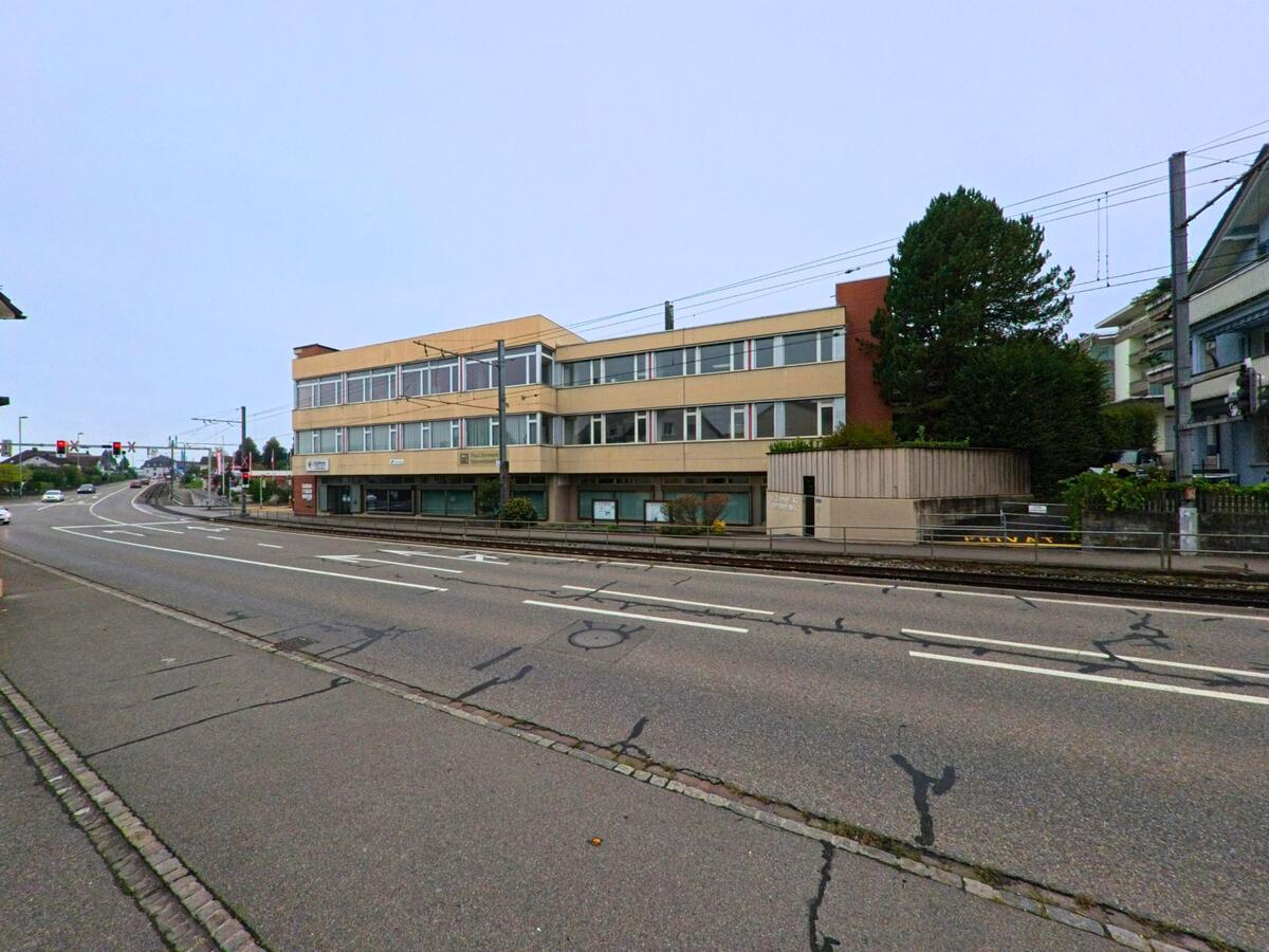 In Unterentfelden (5035, Kanton Aargau) fuehrt eine mehrspurige Strasse mit Tramgleisen an einem laenglichen Verwaltungsgebaeude vorbei.