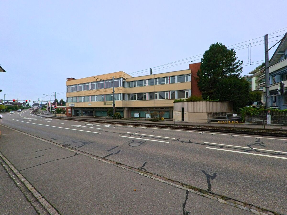 In Unterentfelden (5035, Kanton Aargau) fuehrt eine mehrspurige Strasse mit Tramgleisen an einem laenglichen Verwaltungsgebaeude vorbei.