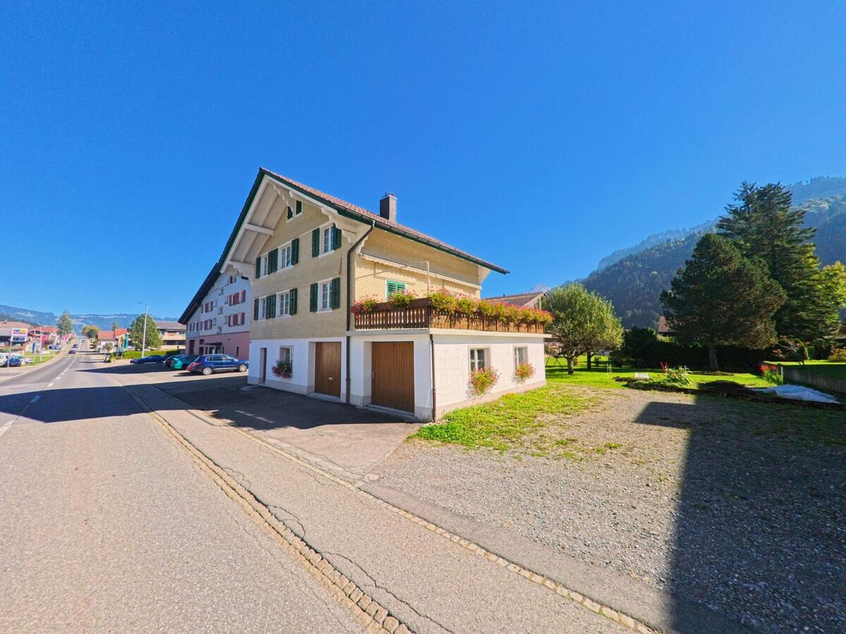 Ein Haus mit gruenen Fensterlaeden steht in Unteriberg (8842, Kanton Schwyz) an einer sonnigen Strasse, waehrend sich rechts eine offene Gruenflaeche vor den bewaldeten Haengen erstreckt.