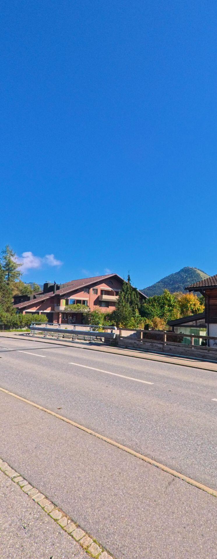 An einer breiten Strasse in Unteriberg (8842, Kanton Schwyz) steht ein Wohnhaus mit dunkler Fassade, waehrend im Hintergrund ein bewaldeter Huegel sichtbar ist.