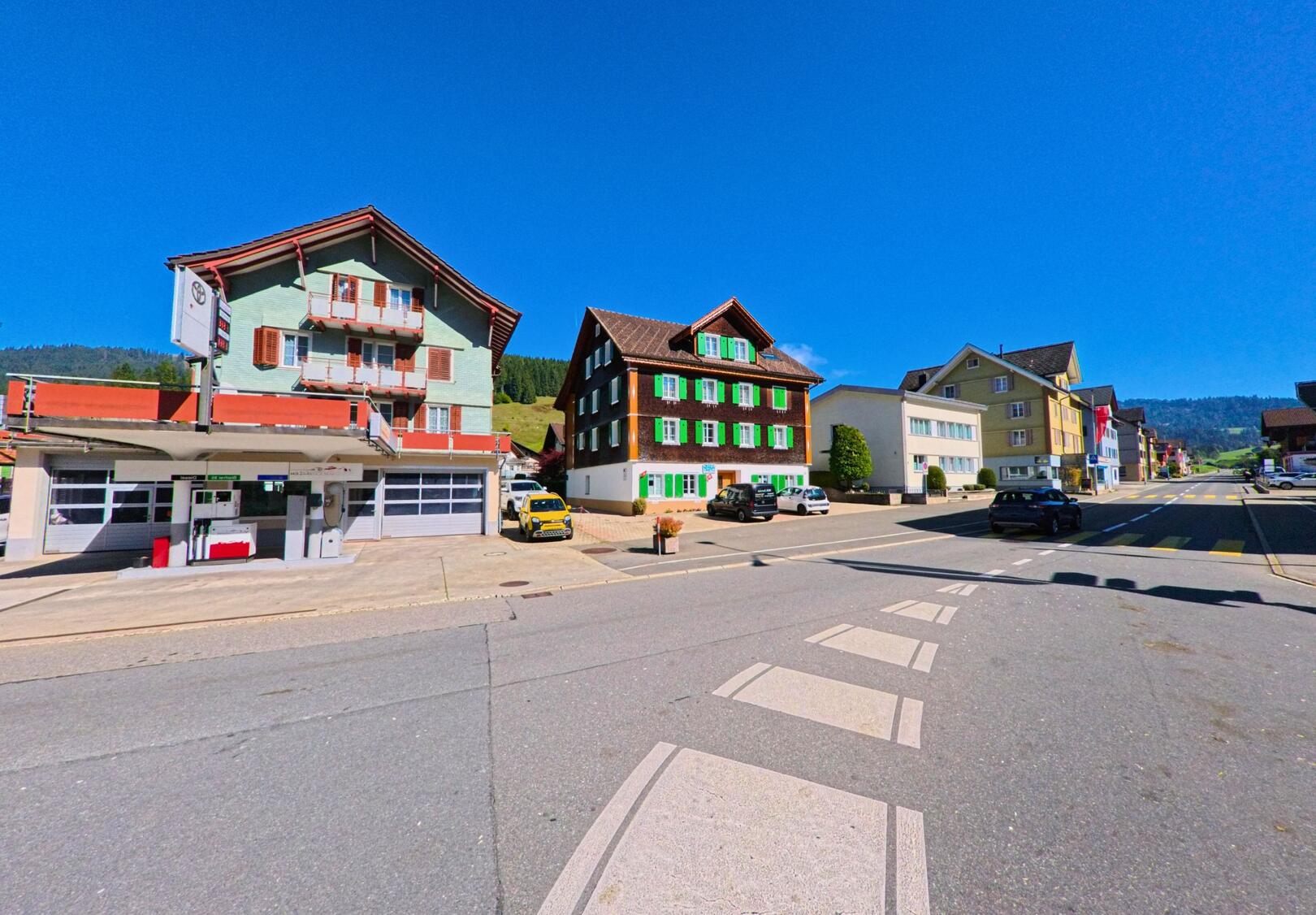 Im Zentrum von Unteriberg (8842, Kanton Schwyz) stehen entlang der Strasse mehrere farbige Wohn- und Gewerbehaeuser, die bei klarem Himmel und Sicht auf die umliegenden Haenge gut erkennbar sind.
