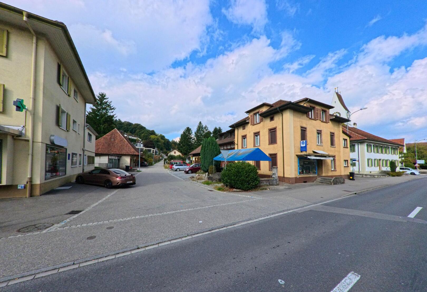 In Unterkulm (5726, Kanton Aargau) zeigt das Bild eine ruhige Strasse mit Wohn- und Gewerbehaeusern sowie parkenden Autos unter einem leicht bewoelkten Himmel.
