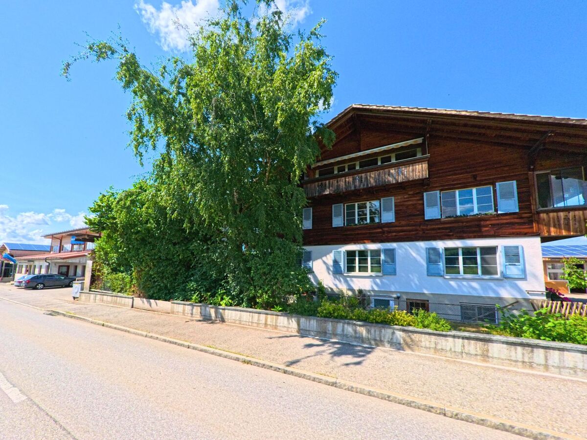 In Unterlangenegg (3614 im Kanton Bern) steht ein Wohnhaus mit Holzfassade und blauen Fensterlaeden hinter einem grossen Baum direkt an der Strasse.